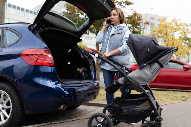 Volwassen moeder met een kinderwagen bij de open kofferbak van een auto spreekt aan de telefoon