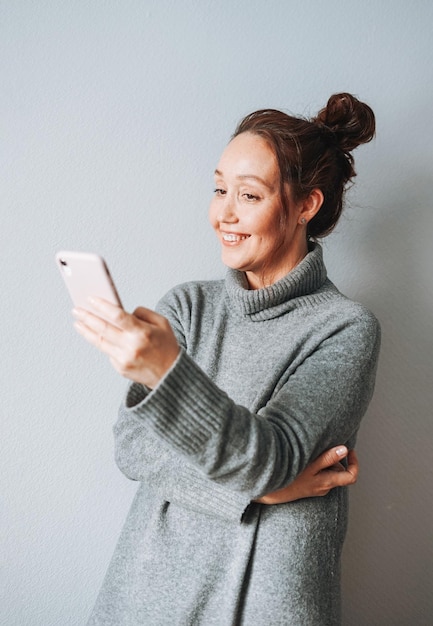 Volwassen mooie lachende vrouw veertig jaar met donkerbruin krullend haar in warme grijze gebreide jurk met mobiel op grijze achtergrond