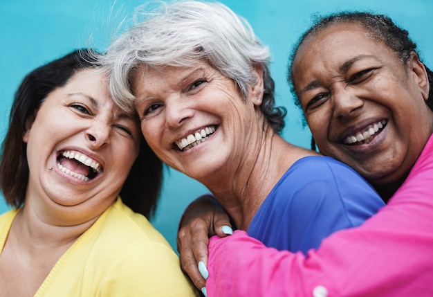 Volwassen multiraciale vrouwen knuffelen elkaar in de stad met blauwe achtergrond terwijl ze lachen op camera - ouderen en vriendschap liefde concept