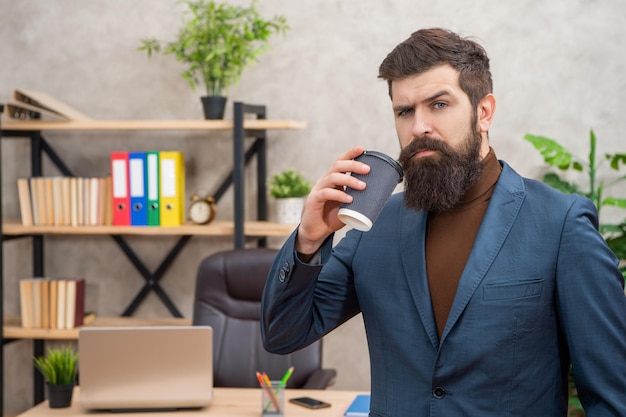 Volwassen ondernemer in jas met koffiepauze met kopje op kantoor afhaalmaaltijden