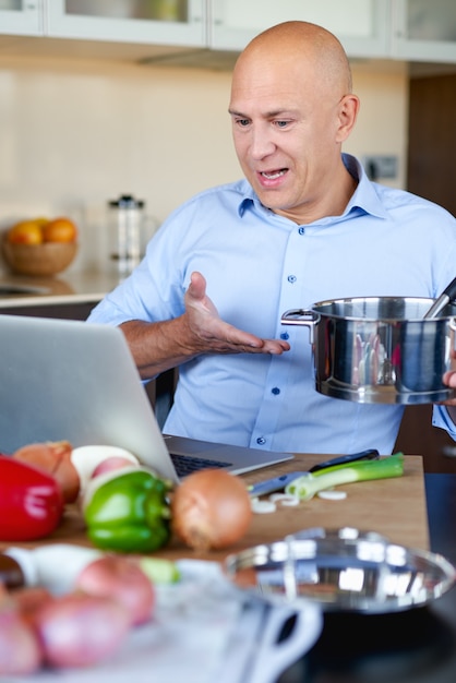 Volwassen sterke man in keuken bereiden van voedsel en onderzoekt laptop