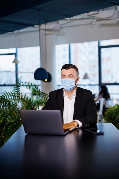 Volwassen succesvolle knappe mannelijke zakenman in een masker met een laptop terwijl hij in een extern kantoor in een café werkt