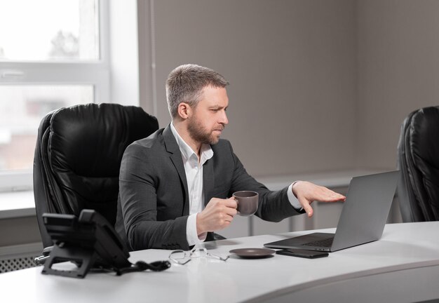 Volwassen succesvolle man in een grijs pak zit aan een bureau met een kopje koffie in zijn hand