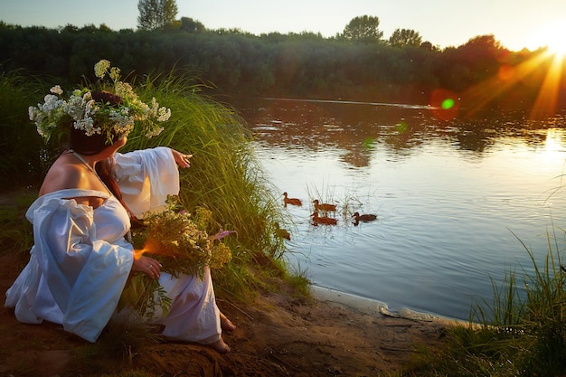 Foto volwassen volwassen brunette vrouw in een witte jurk sundress en een krans van bloemen in de zomer bij het water van de rivier of het meer in de avond bij zonsondergang viering van de slavische heidense feestdag van ivan kupala