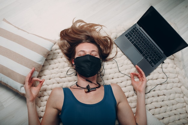 Volwassen volwassen vrouw die yoga nidra doet en in een gezichtsmasker thuis in de woonkamer ligt met online tutorials op laptop.