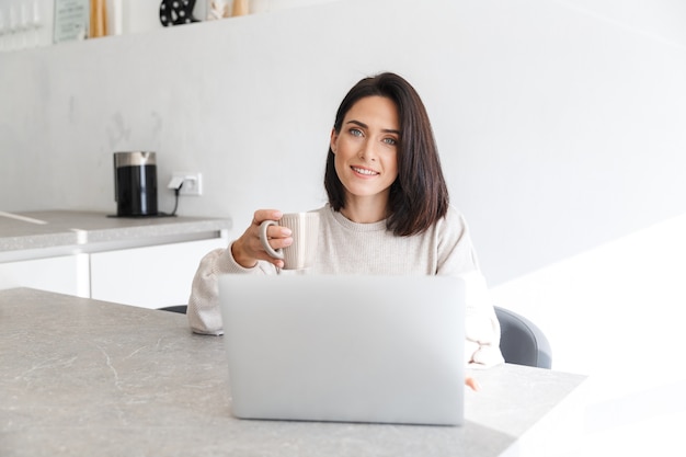 volwassen vrouw 30s die op laptop werkt, zittend over witte muur in lichte kamer