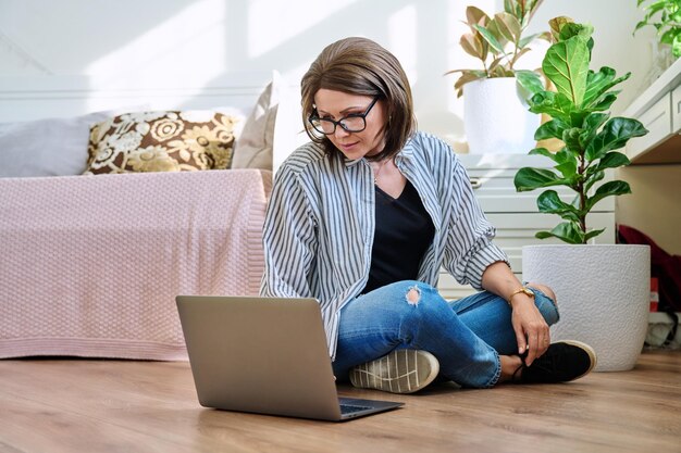Volwassen vrouw die thuis op de vloer zit en naar een laptop kijkt.
