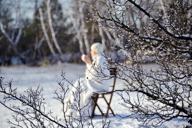 volwassen vrouw in het wit, met mok in zijn handen op een besneeuwde bosachtergrond, heldere zonnige ijzige dag. focus op de voorgrond.