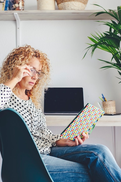 Volwassen vrouw las een boek thuis zittend op de stoel met computer op de achtergrond. indoor moderne mensen vrijetijdsbesteding en ontspannen concept. middelbare leeftijd volwassen student lezen