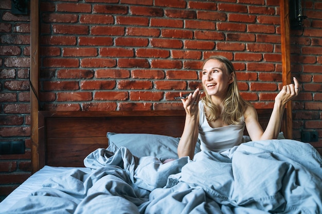 Volwassen vrouw van veertig jaar oud met blond lang haar in pyjama met behulp van mobiele telefoon zittend op bed in zolderkamer thuis