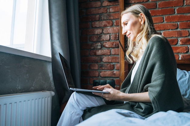 Volwassen vrouw veertig jaar freelancer met blond lang haar in vrijetijdskleding met laptop die thuis op bed zit