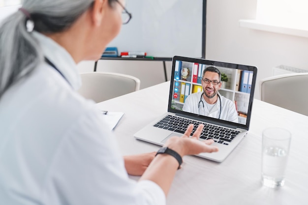 Volwassen vrouwelijke arts heeft videogesprek en chat met collega op laptop in kliniek