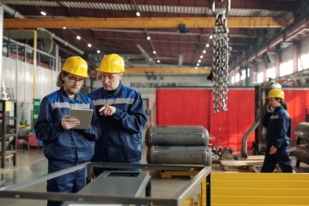 Volwassen werknemer in veiligheidshelm wijzend naar tablet en productieproblemen bespreken met collega terwijl ze bij metalen kubusframe staan