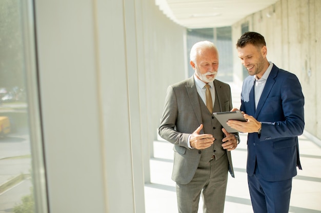 Volwassen zakenman en zijn jonge collega bespreken een financieel verslag op een digitale tablet in de kantoorcorridor