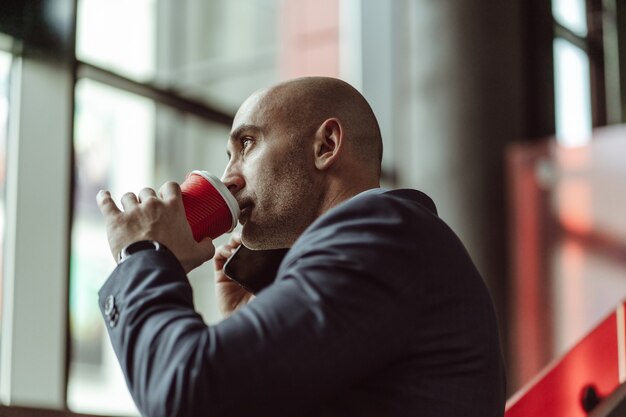 Volwassen zakenman in een pak, praten aan de telefoon tijdens het drinken van koffie met wegwerp