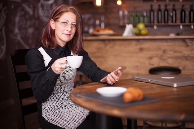 Volwassen zakenvrouw die een smartphone gebruikt terwijl ze een kopje koffie vasthoudt. Ontspannen in een vintage coffeeshop