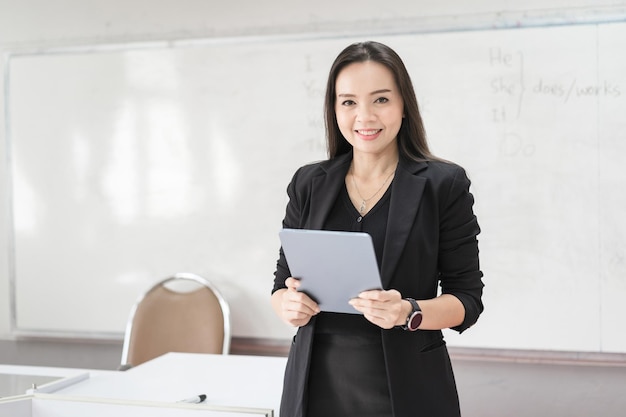 Volwassen zelfverzekerde vrolijke Aziatische vrouwelijke leraar in zwart pak uniform met digitale tablet en laptop die modern lesgeeft in de klas