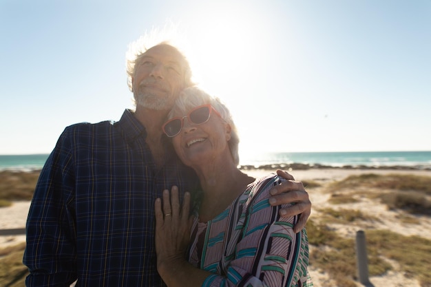 Vooraanzicht close-up van een senior blank stel op het strand in de zon, staand, glimlachend en omhelzend, met de zee op de achtergrond, tegenlicht