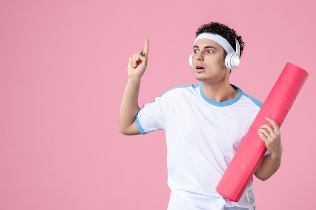 Vooraanzicht jong mannetje in sportkleren met yogamat en hoofdtelefoons op roze muur