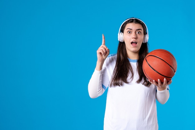 Vooraanzicht jong wijfje dat met oortelefoons basketbal op blauwe muur houdt