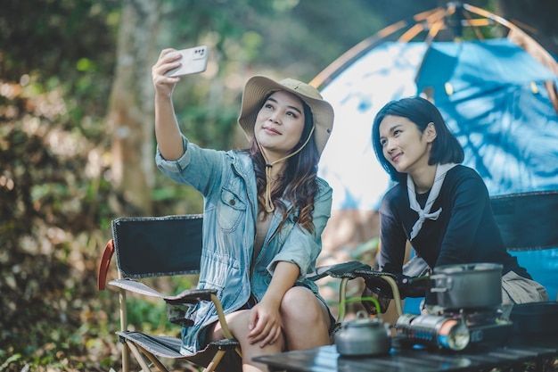 Vooraanzicht Jonge Aziatische mooie vrouw en haar vriendin zitten aan de voorkant van de tent gebruiken mobiele telefoon om foto te maken tijdens het kamperen in het bos met geluk samen