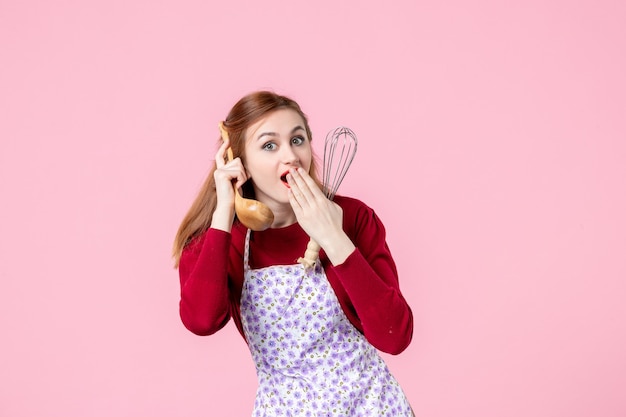 Foto vooraanzicht jonge huisvrouw met garde en houten lepel als telefoon op roze achtergrond keuken beroep horizontale taart vrouw koken uniform cakedeeg