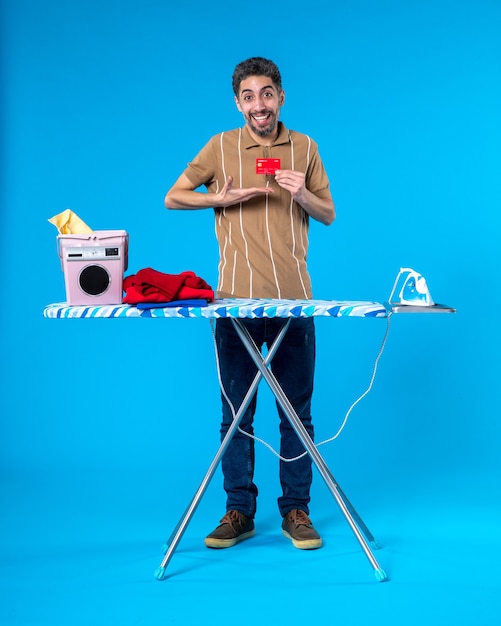 vooraanzicht jonge man achter strijkplank met rode bankkaart op blauwe achtergrond geld schoon huishoudelijk werk wasmachine kleur ijzer
