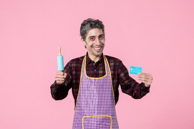 vooraanzicht jonge man in cape met kleine deegroller en bankkaart op roze achtergrond geld beroep echtgenoot uniform koken baan werknemer keuken