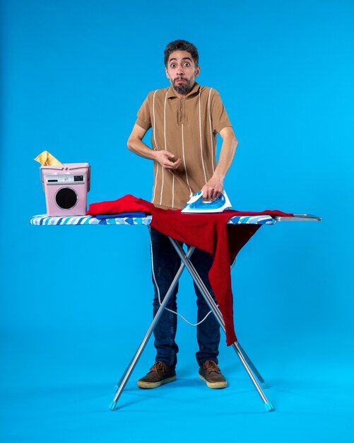 Foto vooraanzicht jonge man strijken achter strijkplank op blauwe achtergrond schone wasgoed wasmachine kleur huishoudelijk werk
