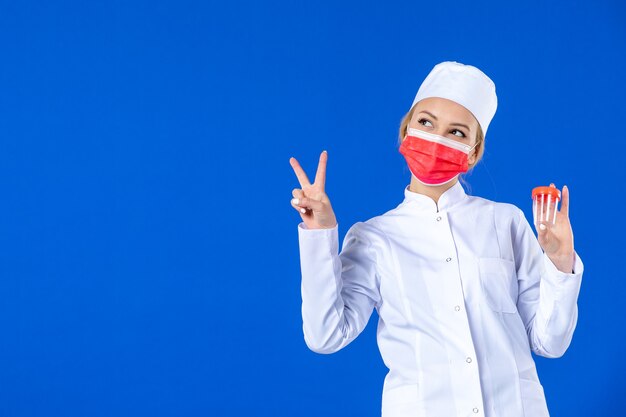 vooraanzicht jonge verpleegster in medisch pak met rood masker met kolven op blauwe achtergrond pandemie covid- ziekenhuis geneeskunde arts gezondheid drug virus