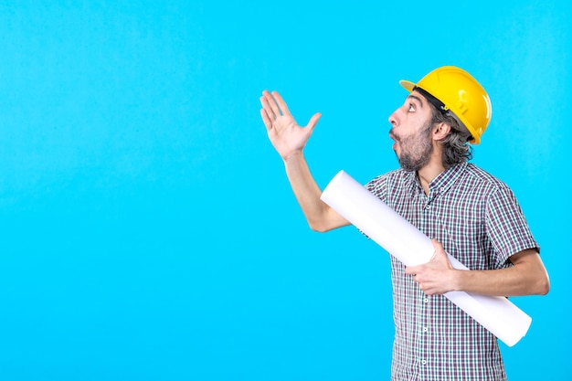 Vooraanzicht mannelijke bouwer in gele helm met plan op een blauwe achtergrond werknemer eigendom architectuur constructeur bouw job engineer