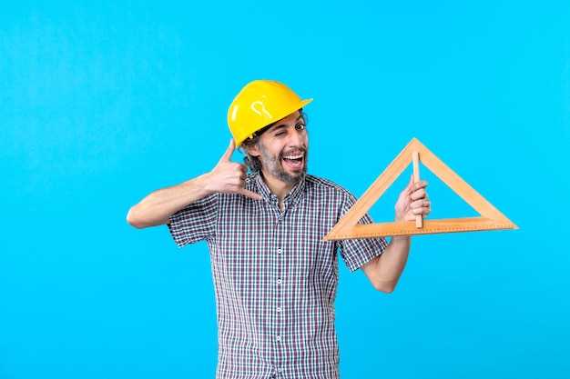 Vooraanzicht mannelijke bouwer in helm met driehoek houten figuur op blauwe achtergrond architectuur baan ingenieur constructor kleur gebouw