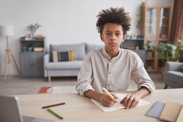 Vooraanzicht portret van Afro-Amerikaanse tienerjongen die huiswerk doet aan het bureau en naar de camera kijkt in het moderne interieur, kopieer ruimte