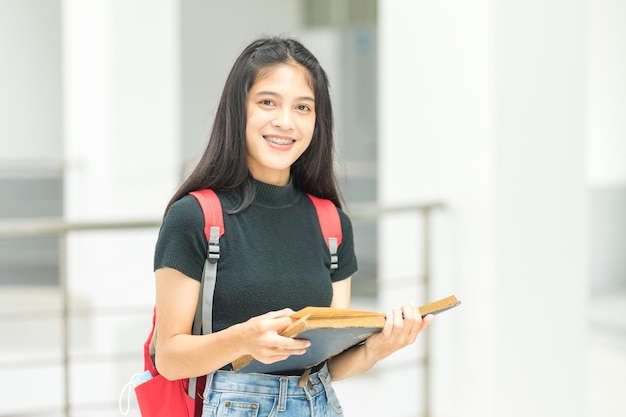 Vooraanzicht portret van een jonge, vrolijke aziatische adolescente studente in een ontspannen, casual terug naar school met boekenstandaards in het campusgebouw