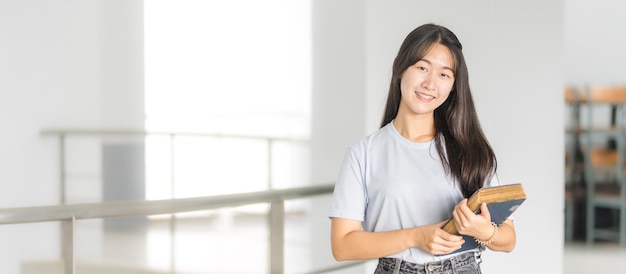 Vooraanzicht portret van een jonge, vrolijke Aziatische adolescente studente in een ontspannen, casual terug naar school met boekenstandaards in het campusgebouw
