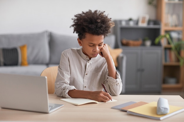 Vooraanzicht portret van tiener Afro-Amerikaanse jongen die huiswerk doet en in notitieboekje schrijft terwijl hij aan een bureau zit met laptop, kopieer ruimte