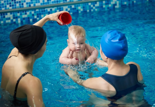 Vooraanzicht van babyjongen tijdens zwemles Achteraanzicht van moeder die kind vasthoudt en vrouw die water op het hoofd van het kind giet