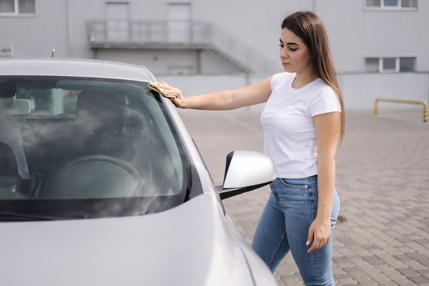 Vooraanzicht van een aantrekkelijke, vrolijke vrouwelijke bestuurder die de koplampen van haar auto wast met een speciale doek erin