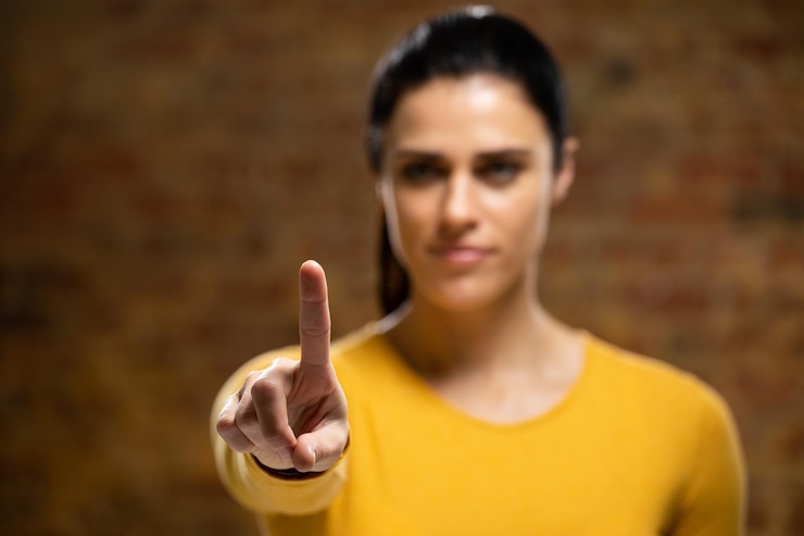 Foto vooraanzicht van een blanke vrouw met lang donker haar, gekleed in een gele top, met opgeheven arm en hand voor haar uitgestoken, met haar wijsvinger naar de camera gericht, terwijl ze een virtueel interactief scherm aanraakt.