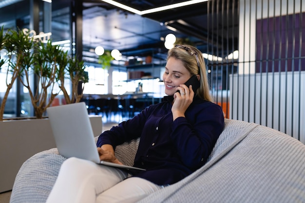 Vooraanzicht van een blanke zakenvrouw die slimme kleding draagt, werkt in een modern kantoor, op een zitzak zit en een laptop en smartphone gebruikt.