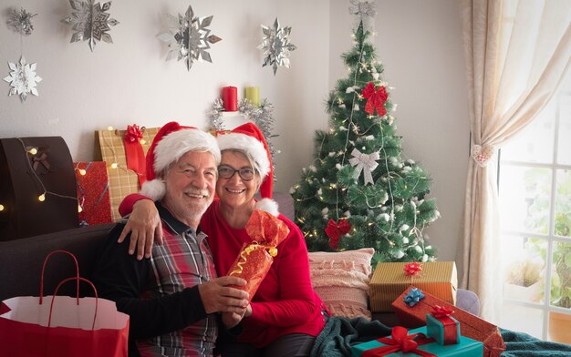 Vooraanzicht van een gelukkig stel senioren die de hoeden van de kerstman dragen en naar de camera kijken. Veel kerstcadeautjes in de buurt voor familie en vrienden.