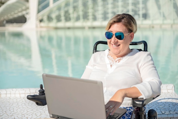 Vooraanzicht van een gelukkige vrouw met zonnebril in rolstoel met behulp van laptop in stedelijke buitenstad scenario