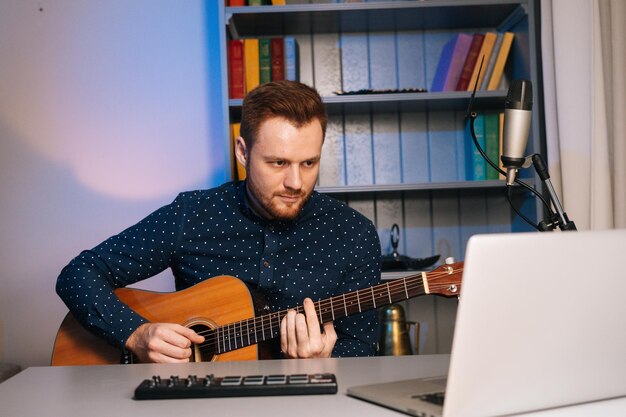 Vooraanzicht van een jonge gitarist die gitaar leert in online muziekles met een verre leraar die aan het bureau zit en op een laptopscherm kijkt