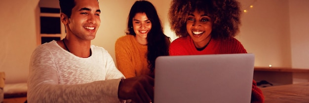 Foto vooraanzicht van een jonge man en vrouw van gemengd ras die aan een tafel zitten met een jonge blanke vrouw die achter hen staat en samen naar een laptopcomputer kijkt en glimlacht in de zitkamer van een appartement