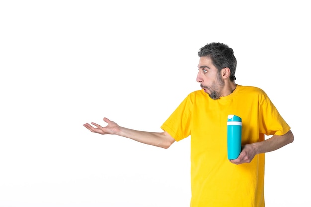 Vooraanzicht van een jonge man in een geel shirt die zich afvraagt en een blauwe thermoskan toont die iets aan de rechterkant op een witte achtergrond wijst