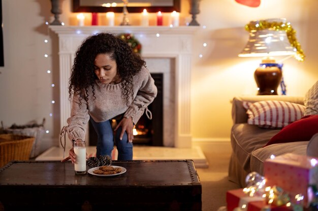 Foto vooraanzicht van een vrouw van gemengd ras in haar zitkamer met kerstmis, die een glas melk naast een bord koekjes op een tafel zet