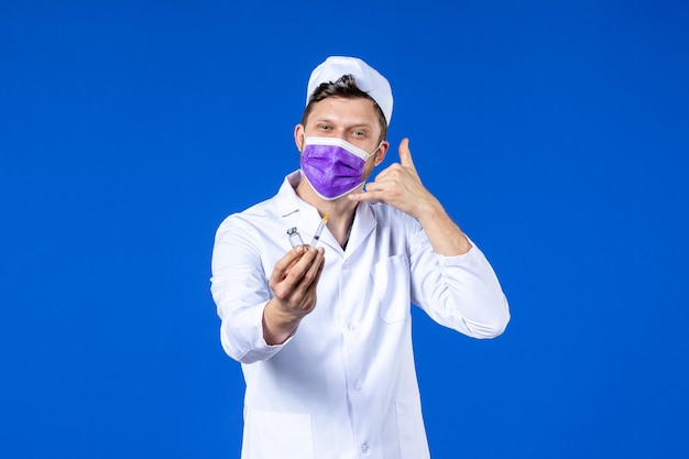 Vooraanzicht van mannelijke arts in medisch kostuum en masker met injectie en vaccin op blauw