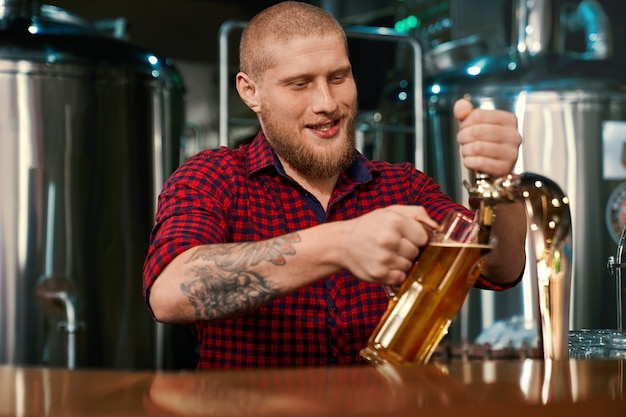 Vooraanzicht van mannelijke barmannen die in pub werken en bier in glas gieten. Jonge, bebaarde man die mensen in brouwerij onderhoudt. Mannetje staat aan de balie, lachen en praten. Concept drank.