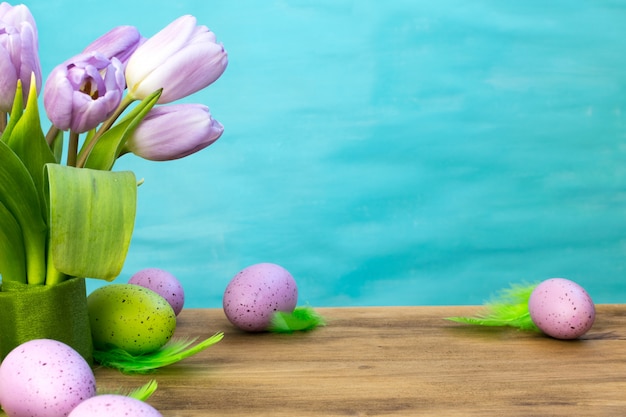 Foto vooraanzicht van paaseieren met groene veren, purpere tulpen op hout en turkooise achtergrond met berichtruimte.