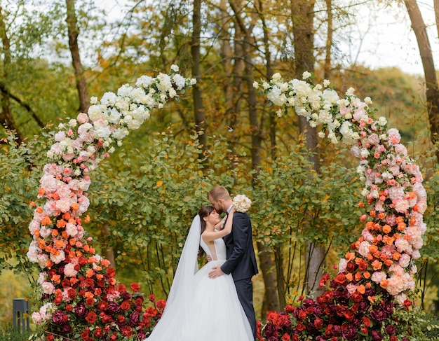 Vooraanzicht van pasgetrouwden knuffelen tijdens een huwelijksceremonie in het park op de achtergrond van een boog van bloemen